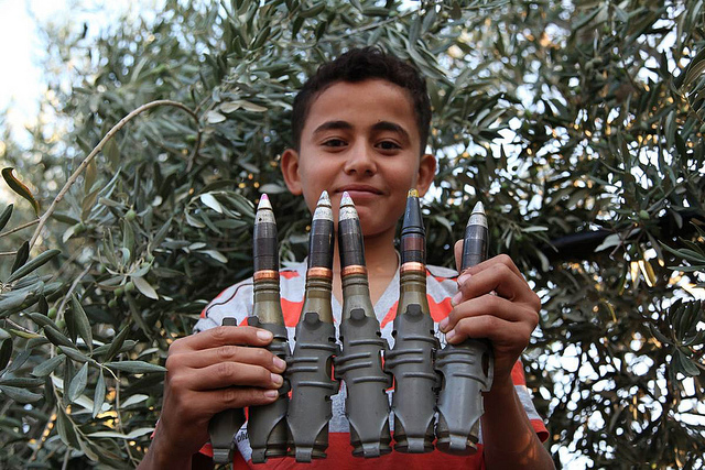 This 2012 photograph shows a Syrian boy holding anti-aircraft rounds up to the camera and smiling in the newly liberated town of Marayan in northern Syria. Photograph by Syria Freedom, shared on flickr and used under (CC BY 2.0